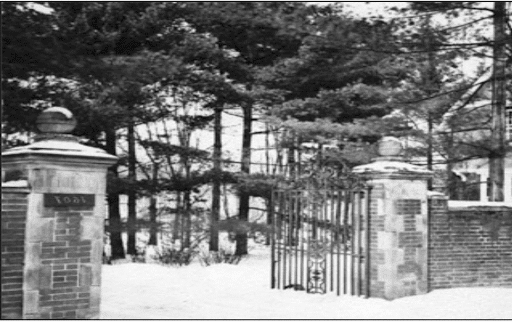 黑白照片，一个锻铁大门，两侧的砖柱与砖墙相连. 树填满了敞开的大门后面的空间.